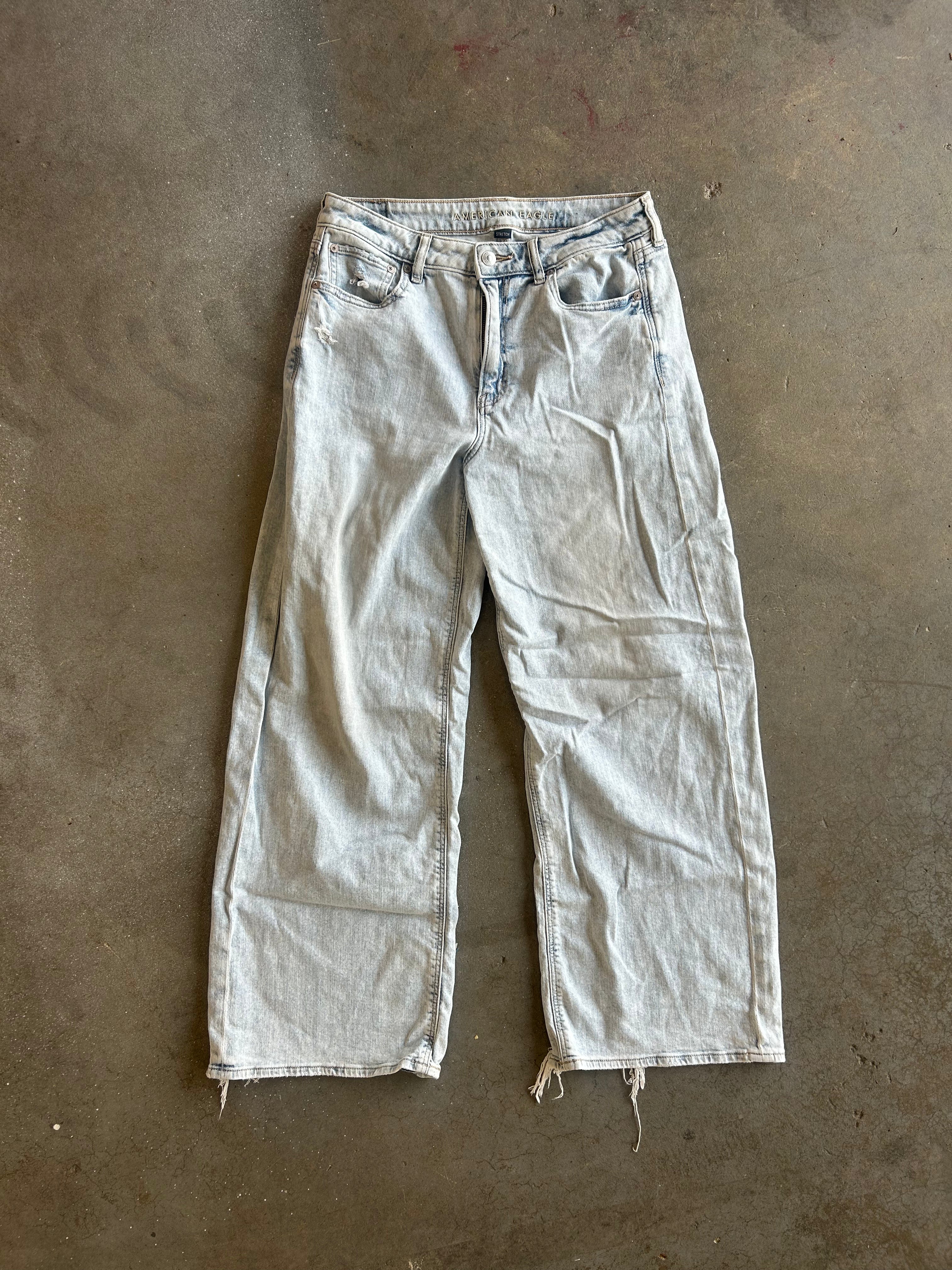 Light-wash faded denim jeans laid flat on a concrete floor, featuring a straight-leg or wide-leg silhouette with raw, frayed hems