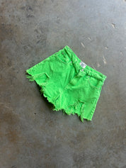Bright neon green denim shorts with a raw, frayed hem, laid flat on a textured gray concrete floor