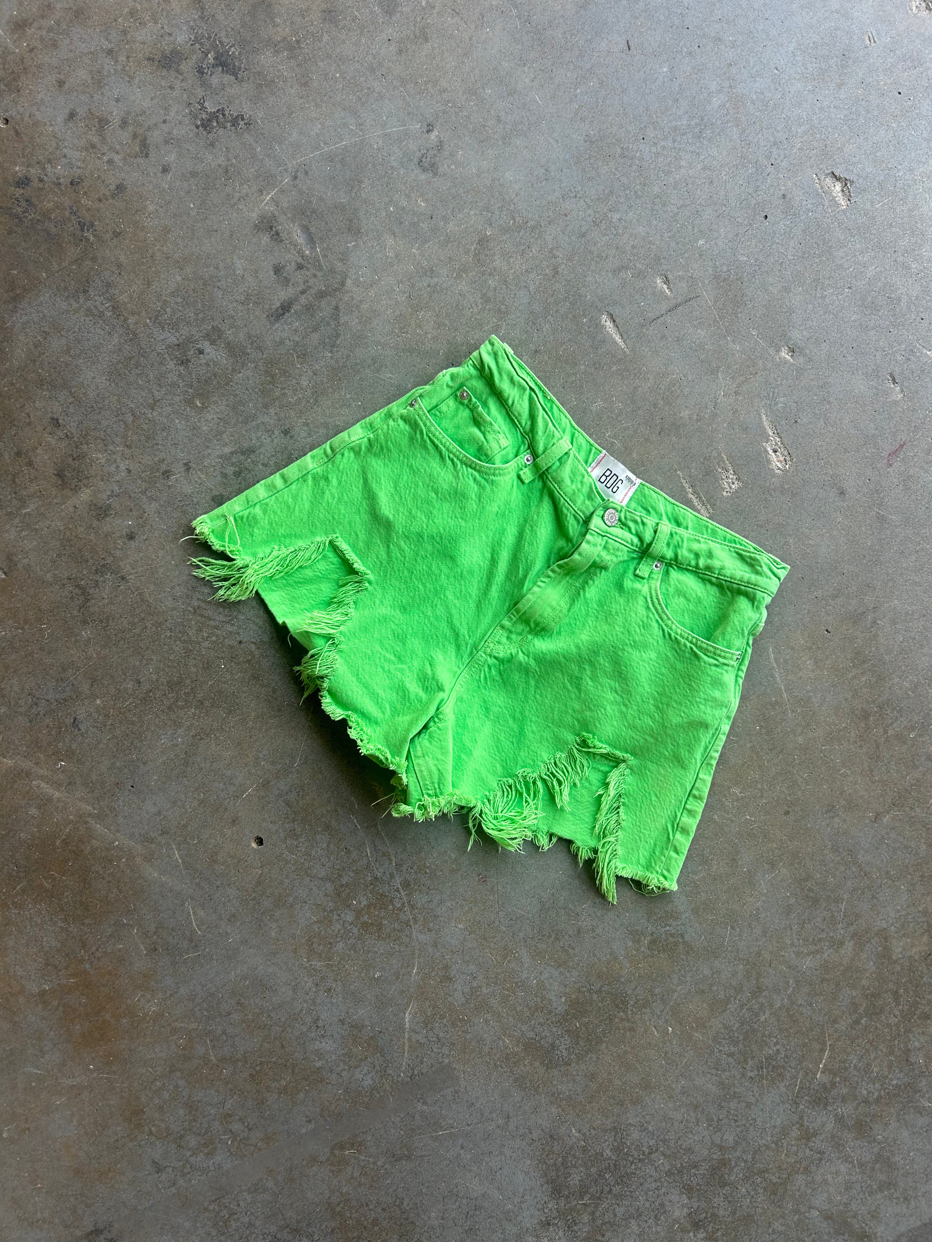 Bright neon green denim shorts with a raw, frayed hem, laid flat on a textured gray concrete floor