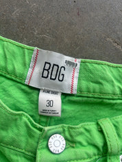 Close-up of bright green denim jeans showing the inner waistband label with 'BDG' branding, size 30, and a metal button closure against a gray concrete background