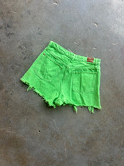 Bright neon green denim shorts with a raw, frayed hem and back pocket details, laid flat on a textured gray concrete floor