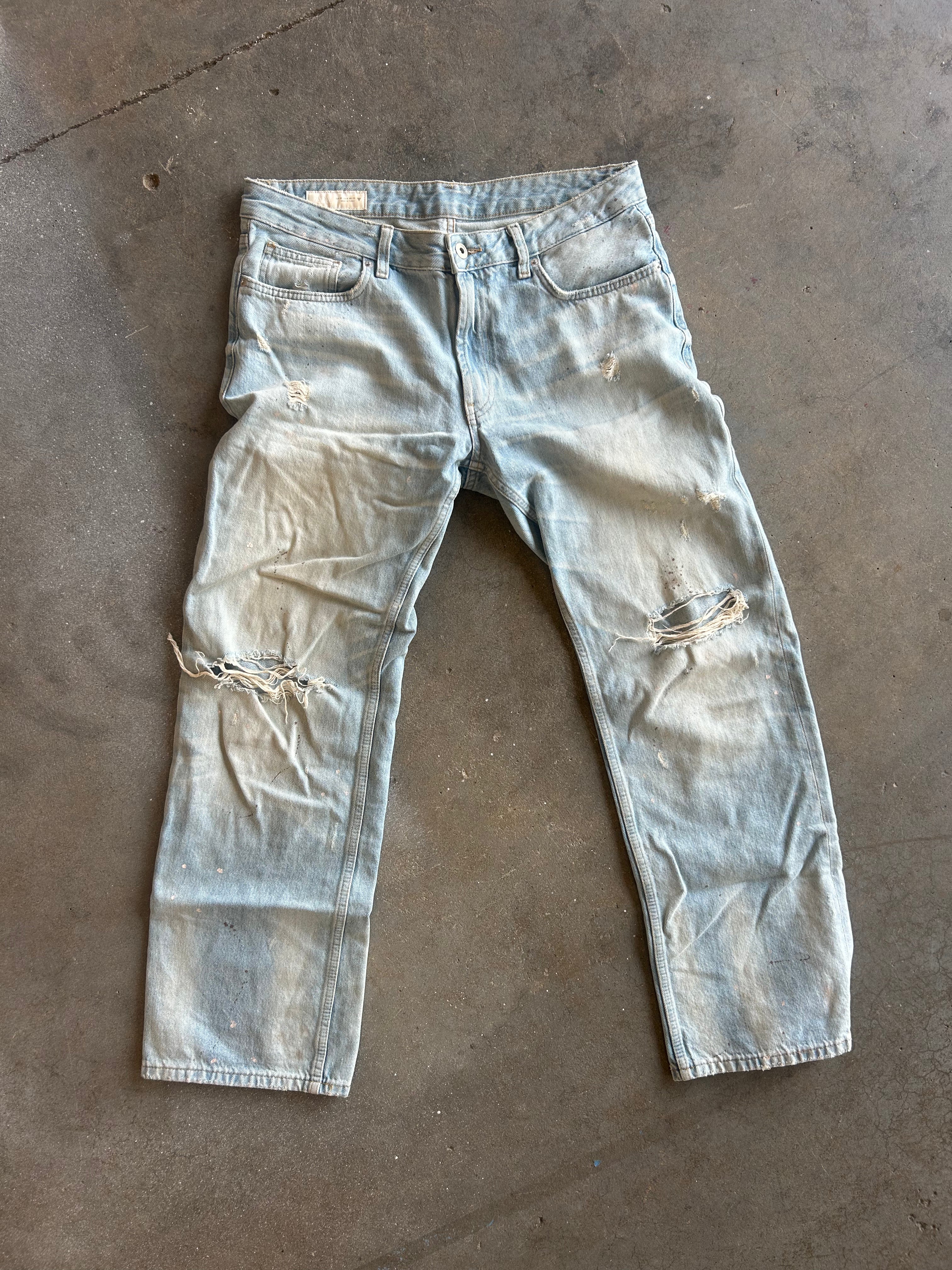 Light-wash distressed denim jeans laid flat on a concrete floor, showing faded blue coloration and ripped details on both legs