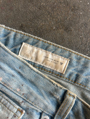 Close-up of light blue denim jeans showing the waistband, stitching, and a beige ZARA brand label sewn inside the waistband, laid on a dark textured surface