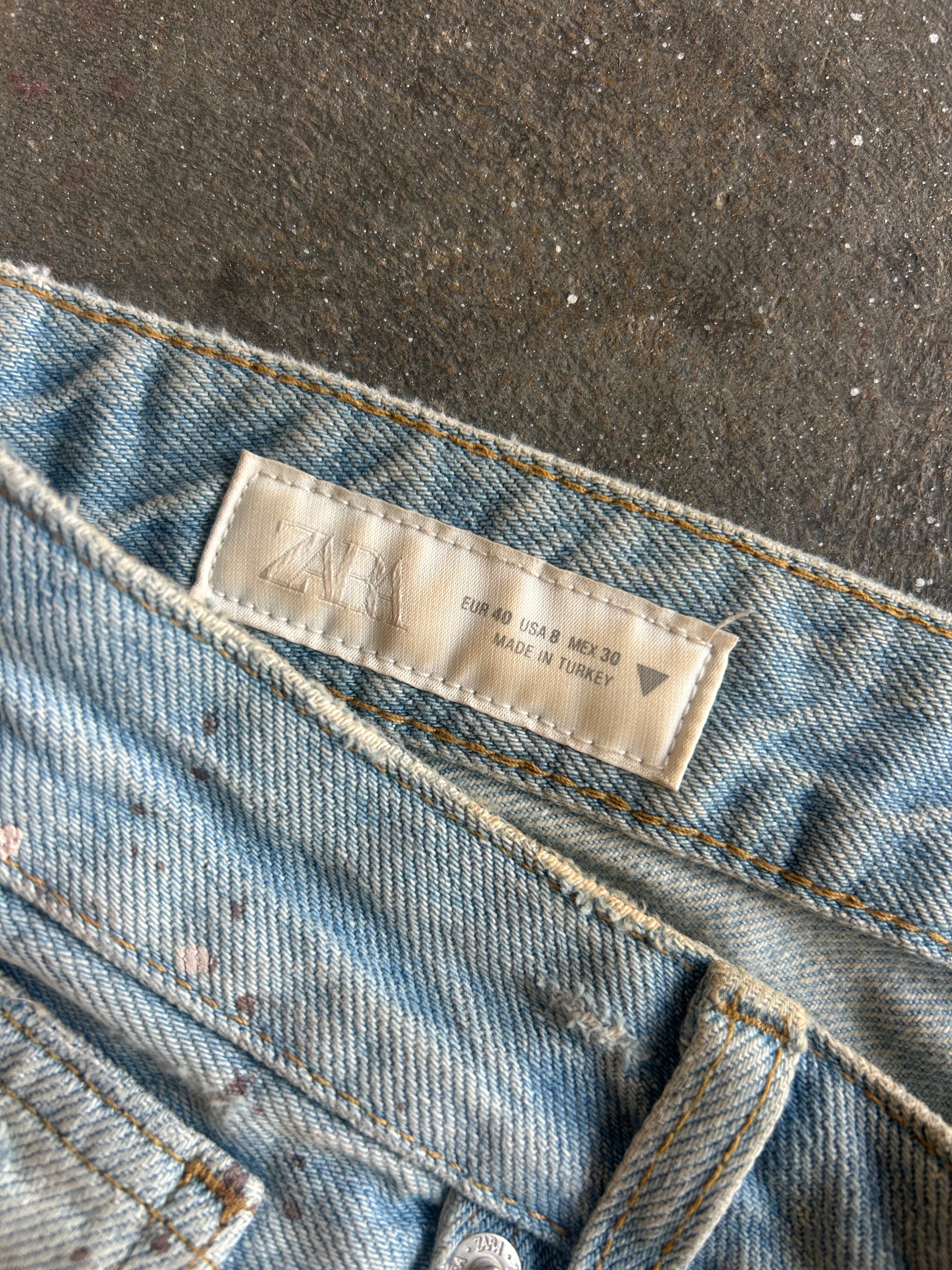Close-up of light blue denim jeans showing the waistband, stitching, and a beige ZARA brand label sewn inside the waistband, laid on a dark textured surface
