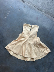 A light beige sleeveless dress or tunic with a gathered, flared silhouette, laid flat on a dark gray concrete surface