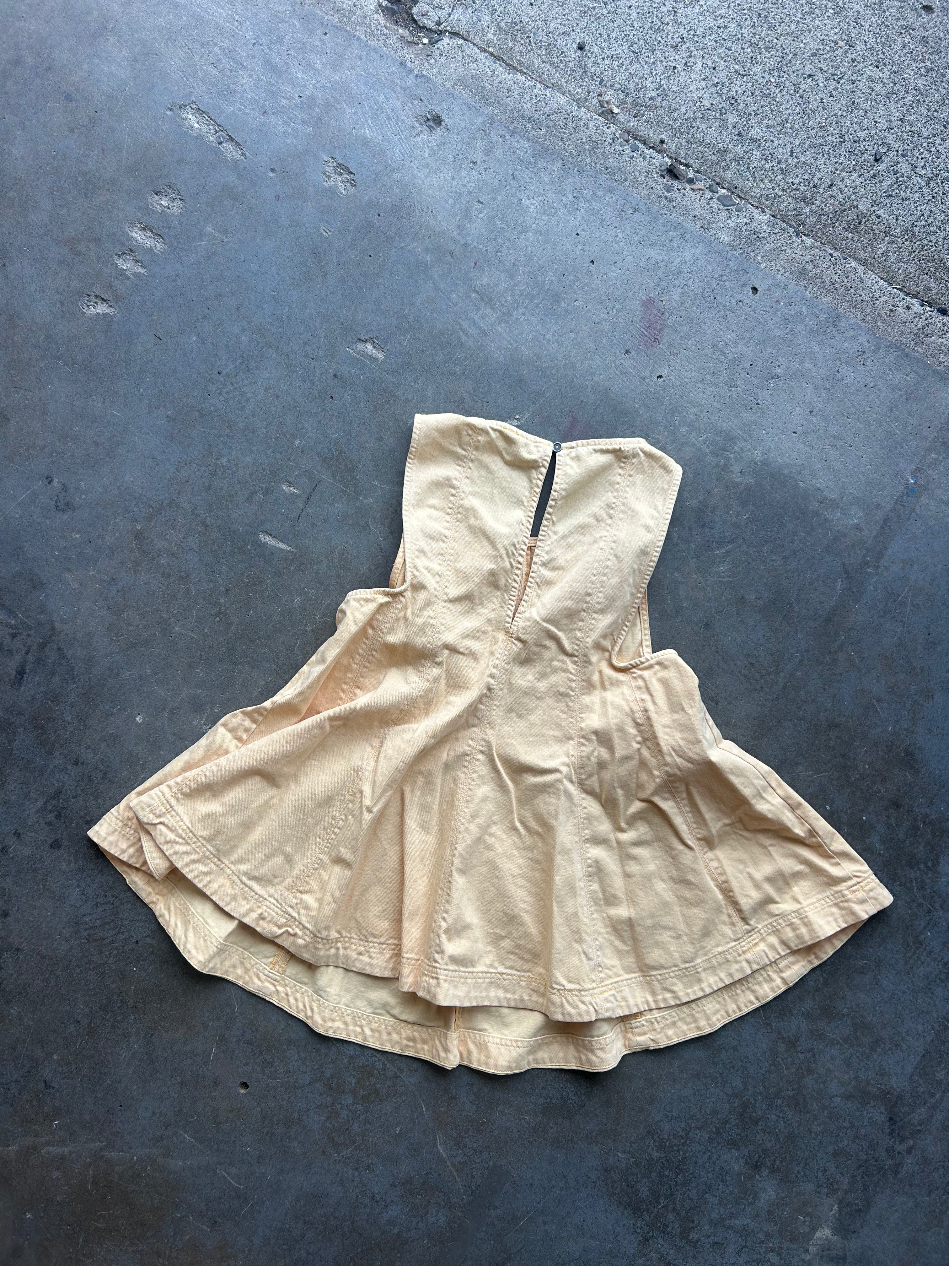 A light beige sleeveless dress or tunic with a gathered, flared silhouette, laid flat on a dark gray concrete surface