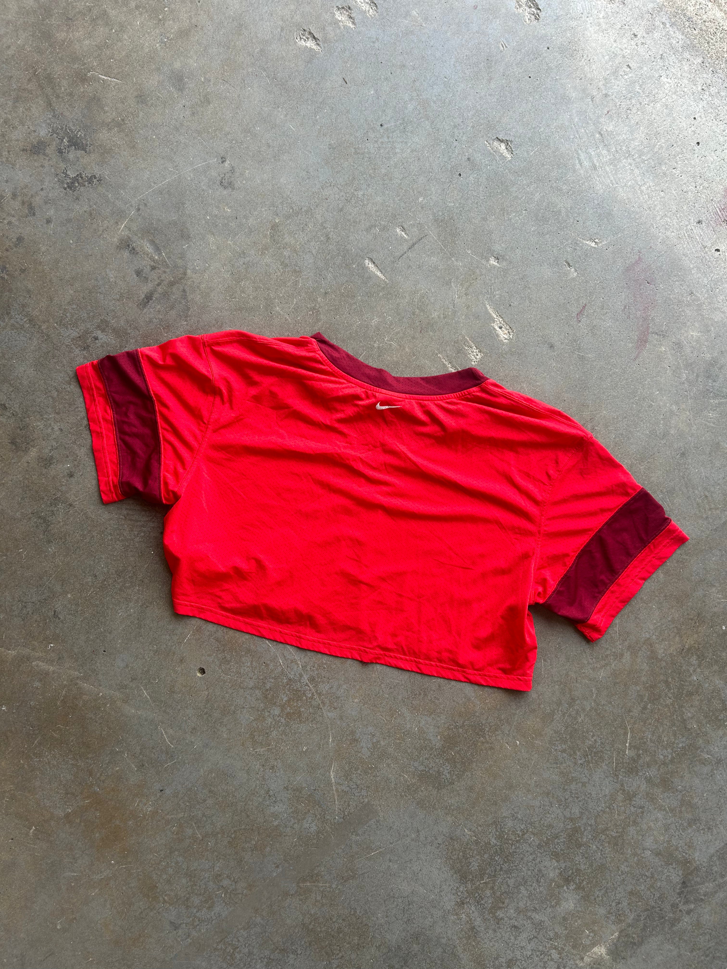 A red short-sleeve crop top with contrasting dark red or burgundy sleeve cuffs and collar, laid flat on a textured gray concrete floor