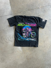 A black vintage Honda motocross t-shirt laid flat on a concrete floor, featuring a colorful graphic of a dirt bike rider and the text 'HONDA motocross WORLD CHAMPIONS'