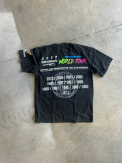 A black short-sleeve Honda World Tour commemorative t-shirt laid flat on a gray concrete floor. The shirt features a large white text list of championship years on the back, with 'WORLD TOUR' printed in vibrant pink, green, and blue letters