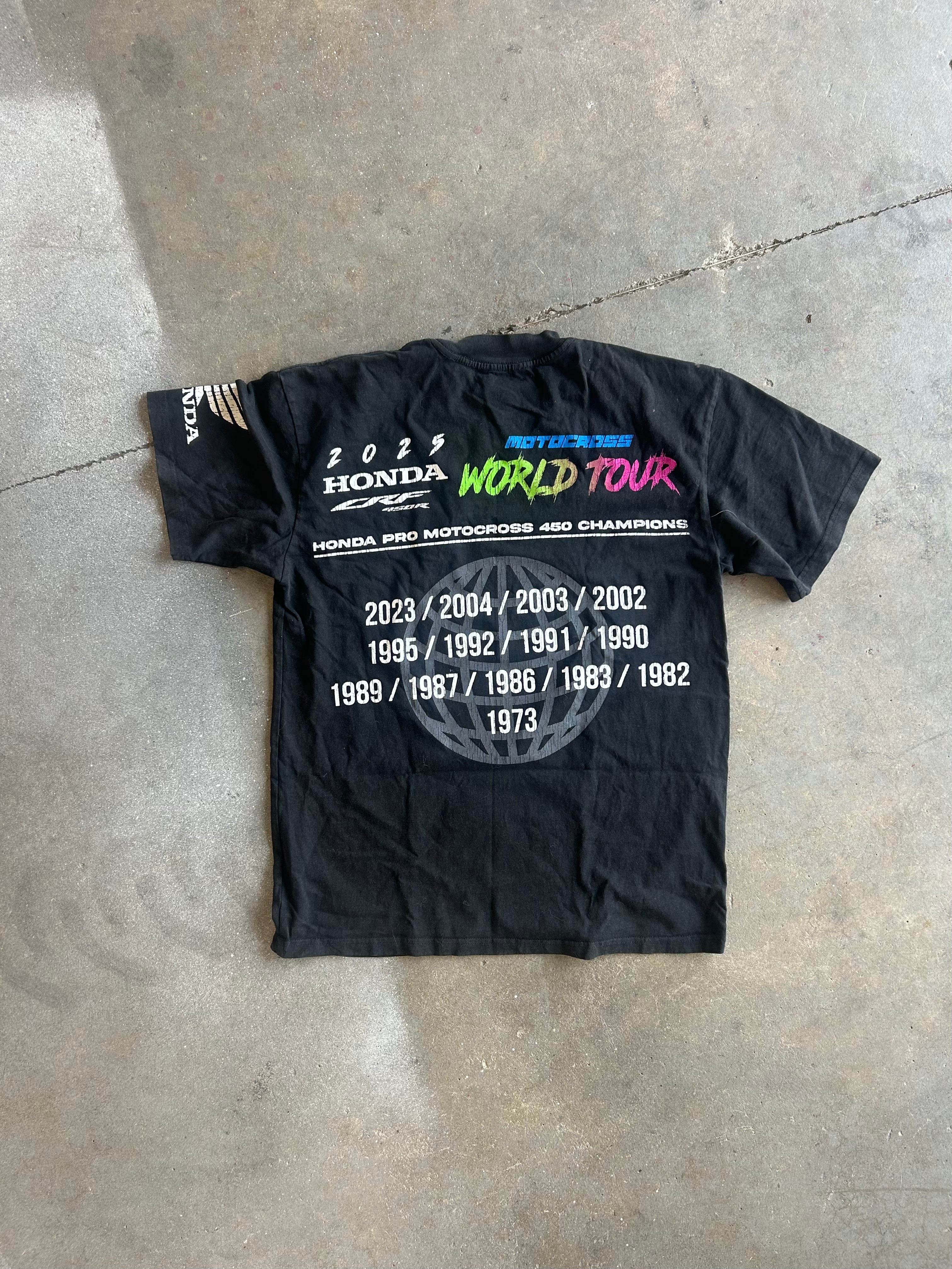 A black short-sleeve Honda World Tour commemorative t-shirt laid flat on a gray concrete floor. The shirt features a large white text list of championship years on the back, with 'WORLD TOUR' printed in vibrant pink, green, and blue letters
