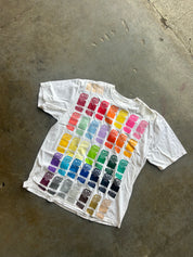 A white short-sleeve t-shirt laid flat on a concrete floor, featuring a large printed grid of colorful paint swatches on the front