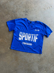 A bright blue cropped V-neck t-shirt with 'SPORTIF' and 'FORWARD' printed in white on the front, laid flat on a concrete floor