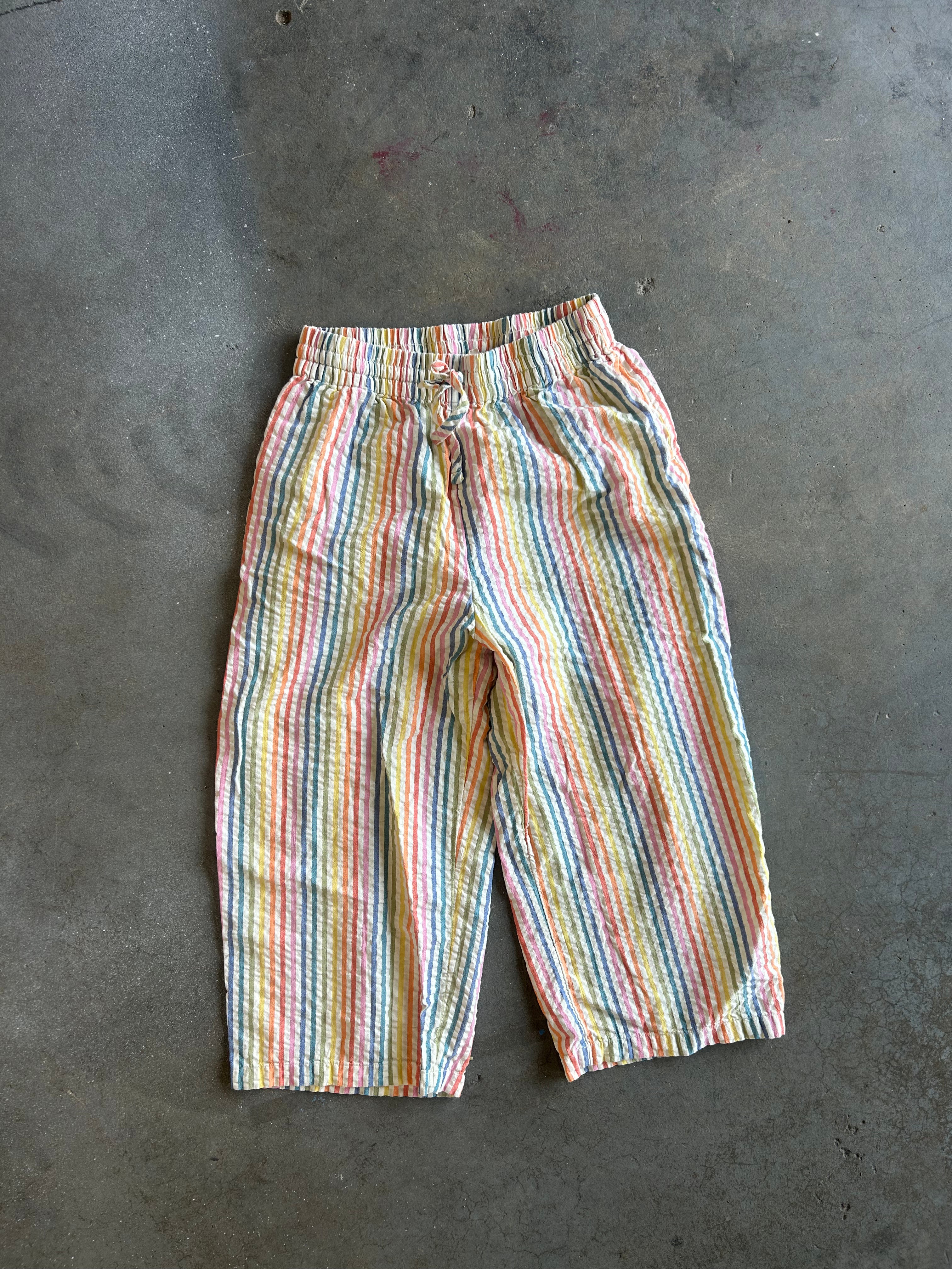 A pair of pastel rainbow-striped cropped pants laid flat on a gray concrete floor