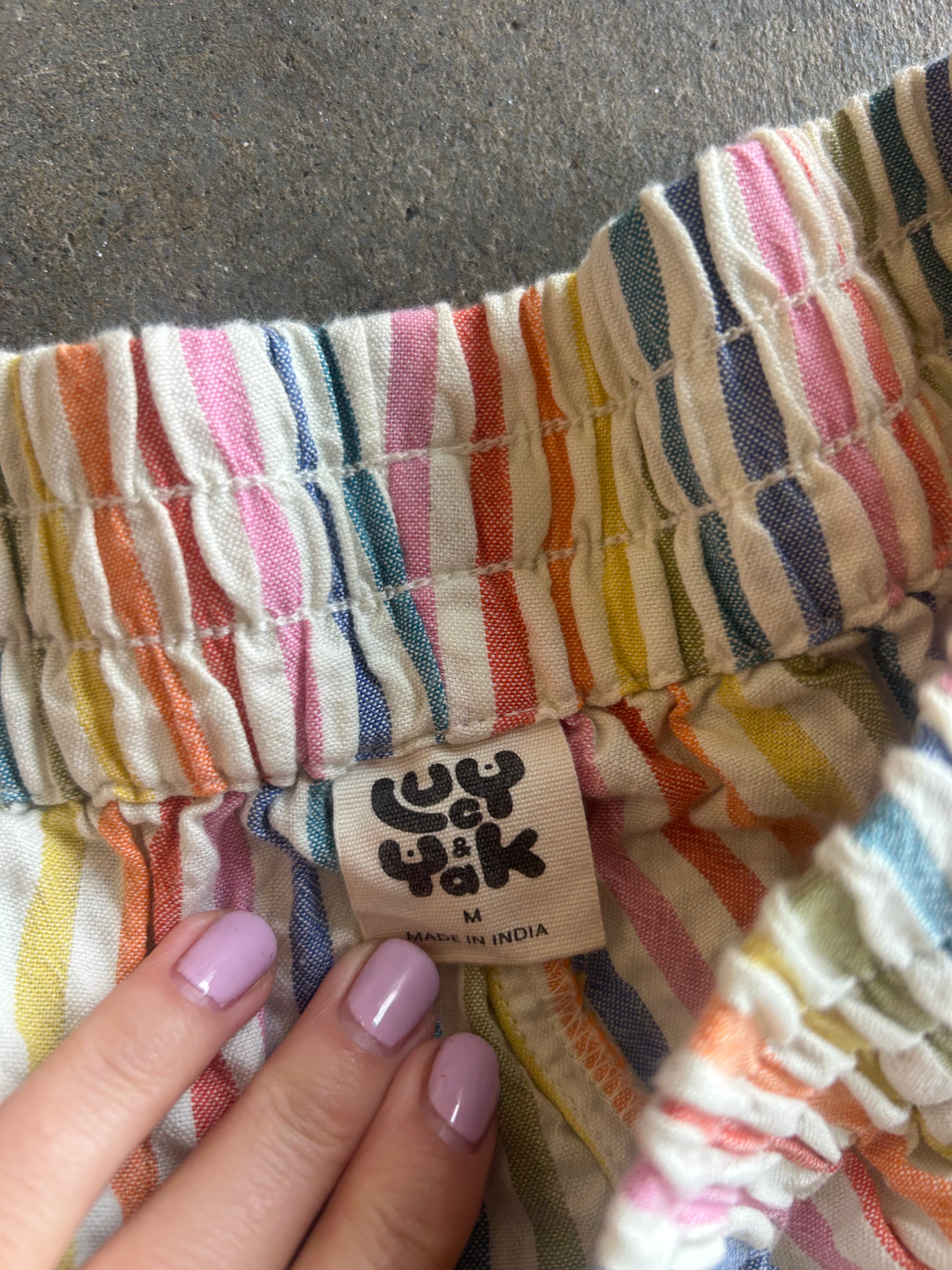 A close-up of a striped garment with a visible fabric tag reading 'LUCY YAK' and 'MADE IN INDIA'. A hand with light purple nail polish is holding the waistband area