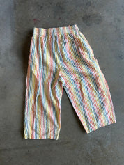 A pair of cropped pants with vertical rainbow stripes in pink, blue, yellow, and green, laid flat on a dark gray concrete floor