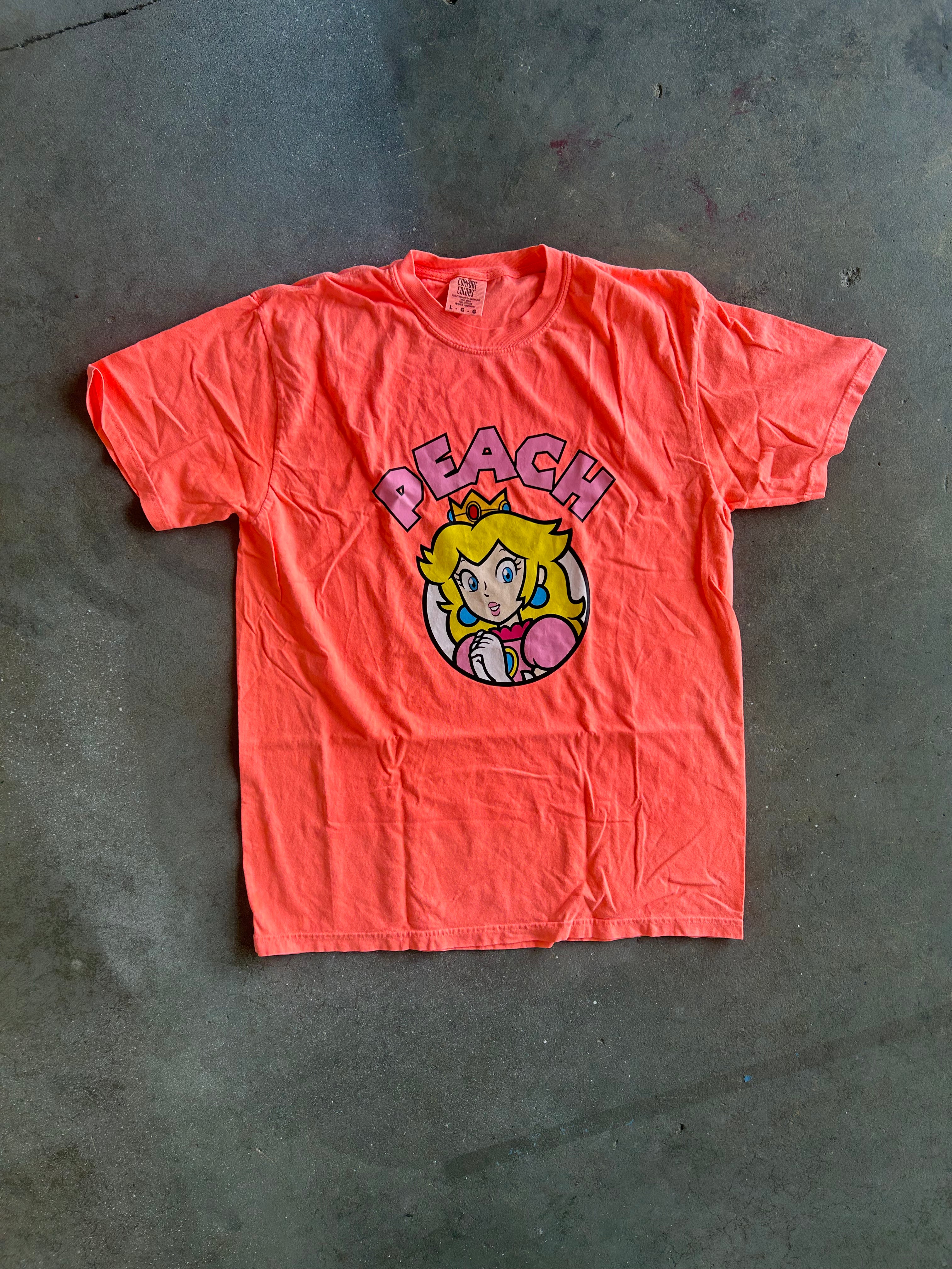 A bright coral pink short-sleeve t-shirt featuring a graphic of the character Peach with the word 'PEACH' above her, laid flat on a gray concrete floor