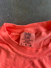 Close-up of a coral pink Comfort Colors t-shirt showing the inner neck label and ribbed collar, laid flat on a gray textured surface