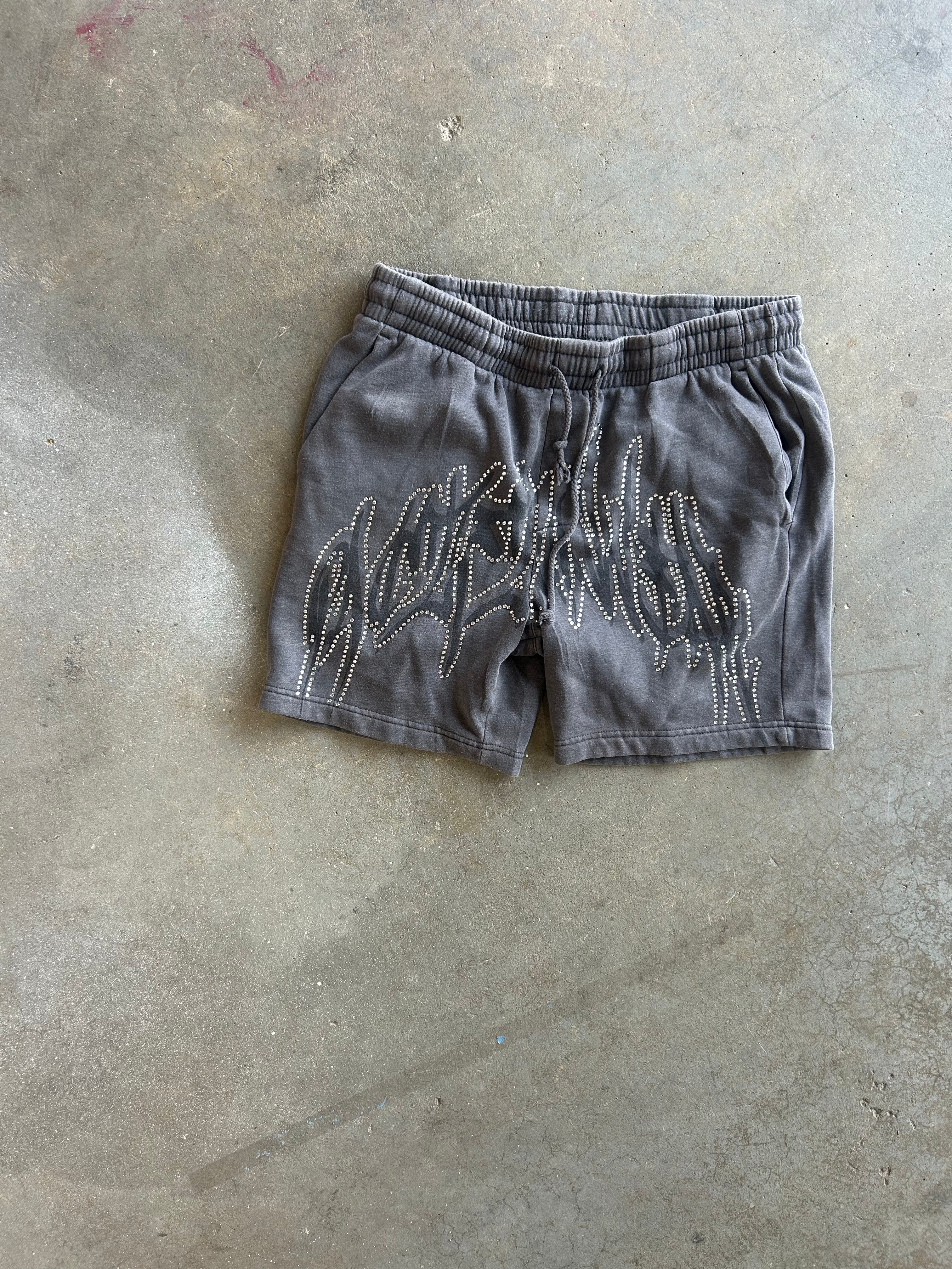 A pair of dark gray sweat shorts with a white graphic design on the front, laid flat on a concrete floor