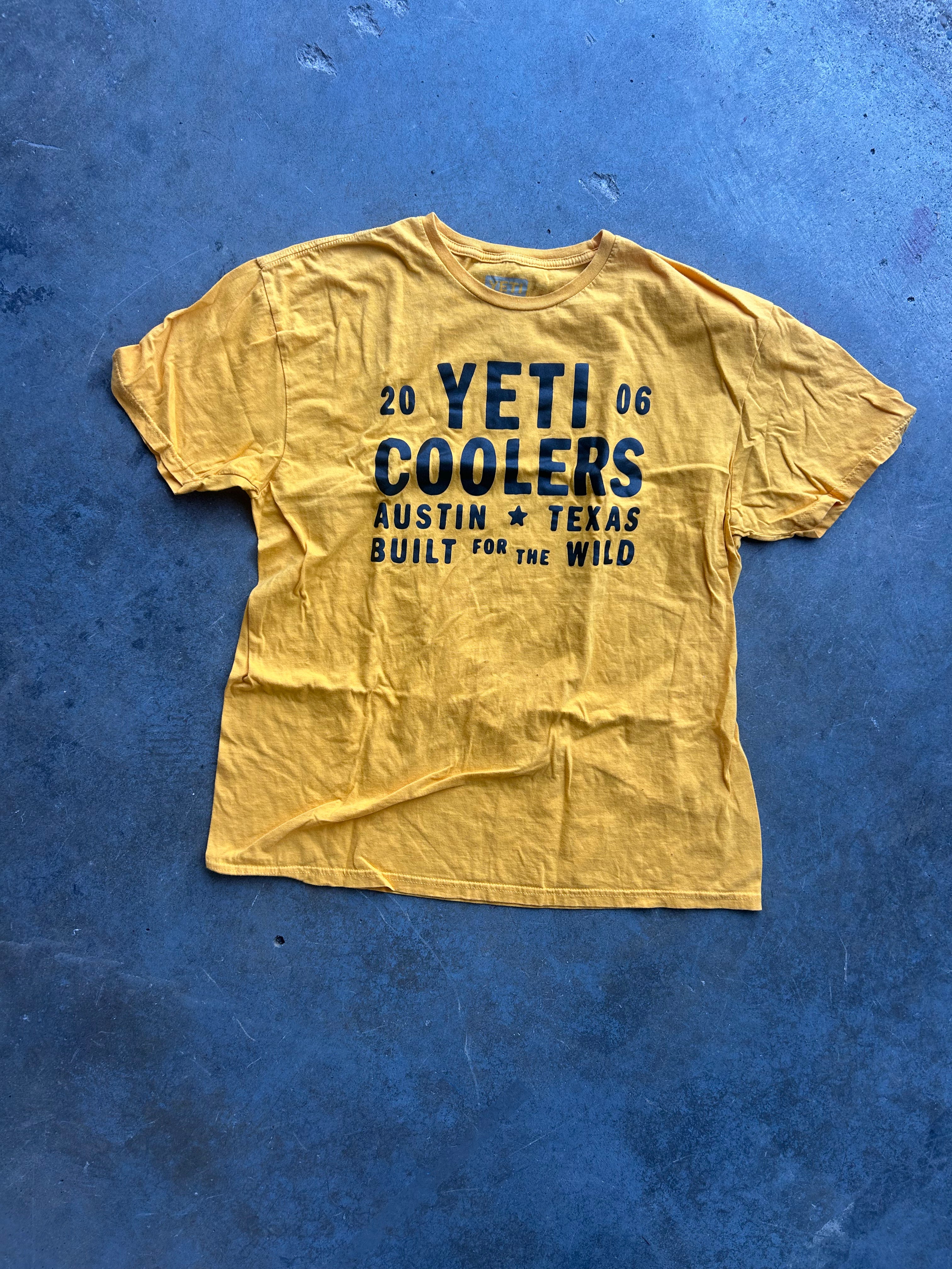 Mustard graphic tee with Yeti Coolers text in navy blue on a concrete surface