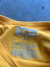 Close-up of a yellow t-shirt collar showing the light blue YETI brand logo and care instructions printed inside the neck, laid flat on a textured blue surface