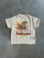 A vintage Budweiser graphic t-shirt in a faded beige color, featuring a running horse and the Budweiser logo, laid flat on a gray concrete floor