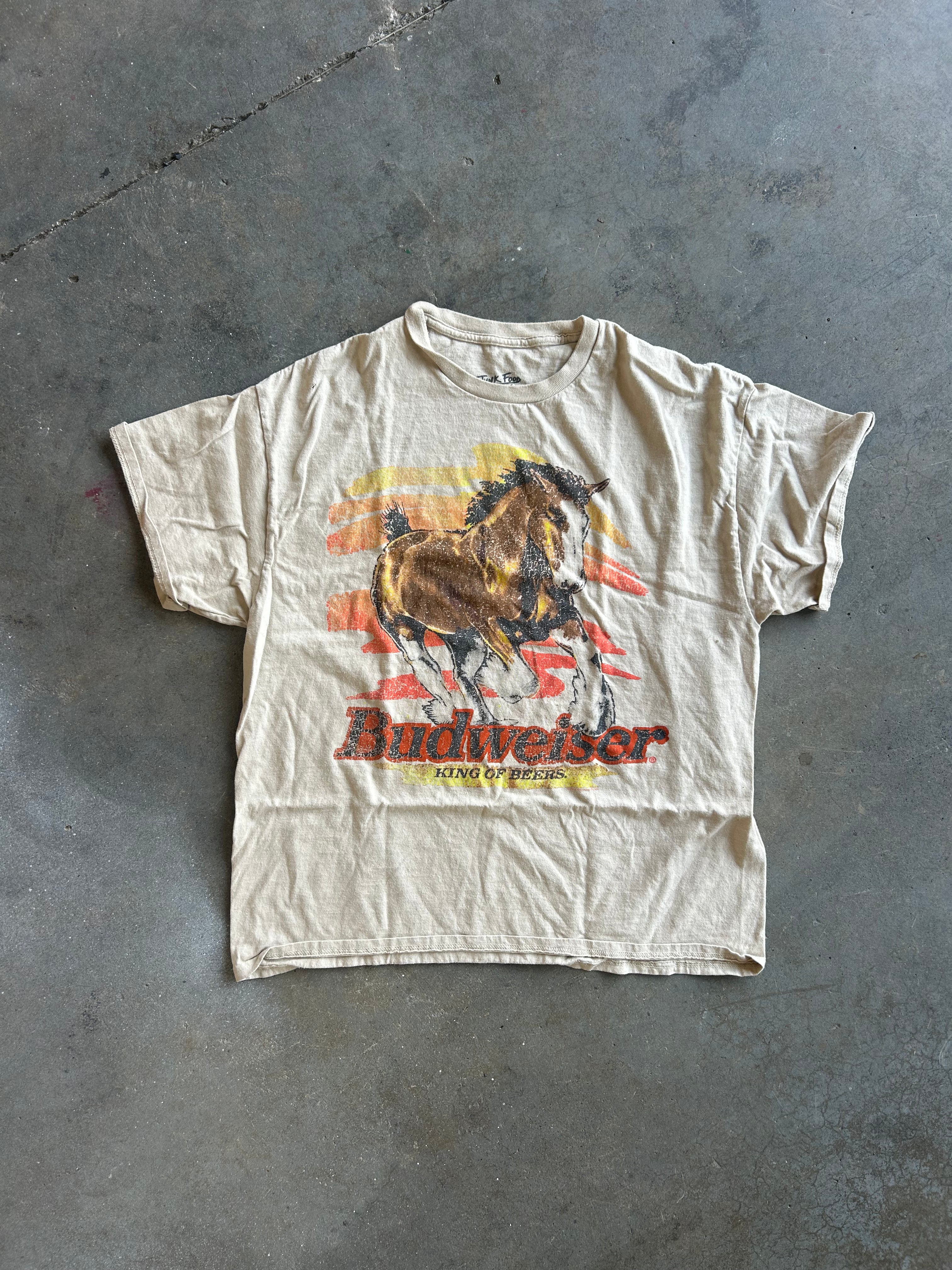 A vintage Budweiser graphic t-shirt in a faded beige color, featuring a running horse and the Budweiser logo, laid flat on a gray concrete floor