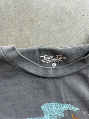 Close-up of the neck collar of a dark gray heather t-shirt laid flat on a concrete surface. The inside collar features a printed brand label reading 'Junk Food Clothing' with a size indicator 'L'