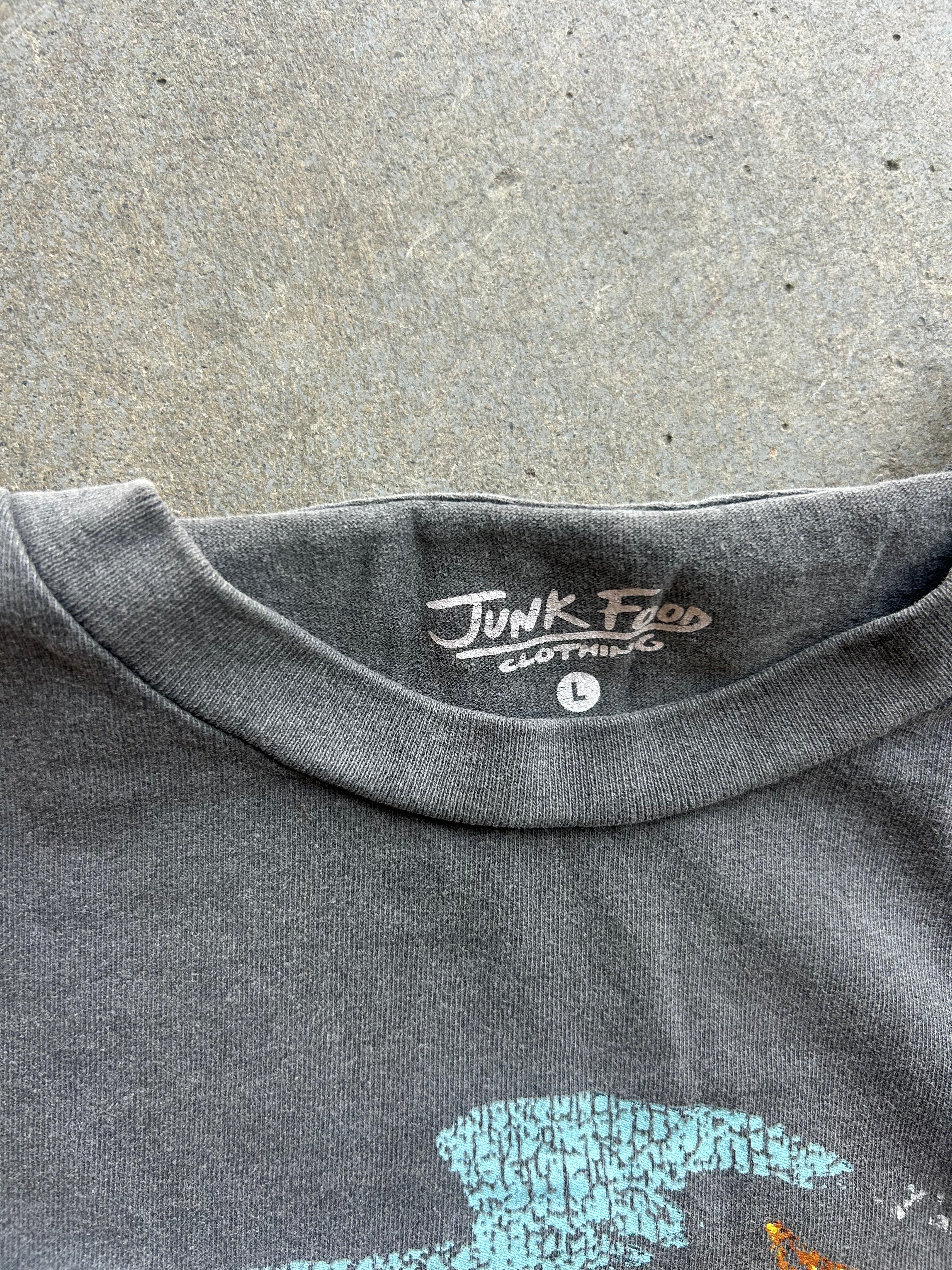 Close-up of the neck collar of a dark gray heather t-shirt laid flat on a concrete surface. The inside collar features a printed brand label reading 'Junk Food Clothing' with a size indicator 'L'