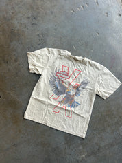 A vintage off-white short-sleeve t-shirt with a distressed graphic of a bird in flight over a red 'X' and the words 'Harley Davidson', laid flat on a textured gray concrete floor