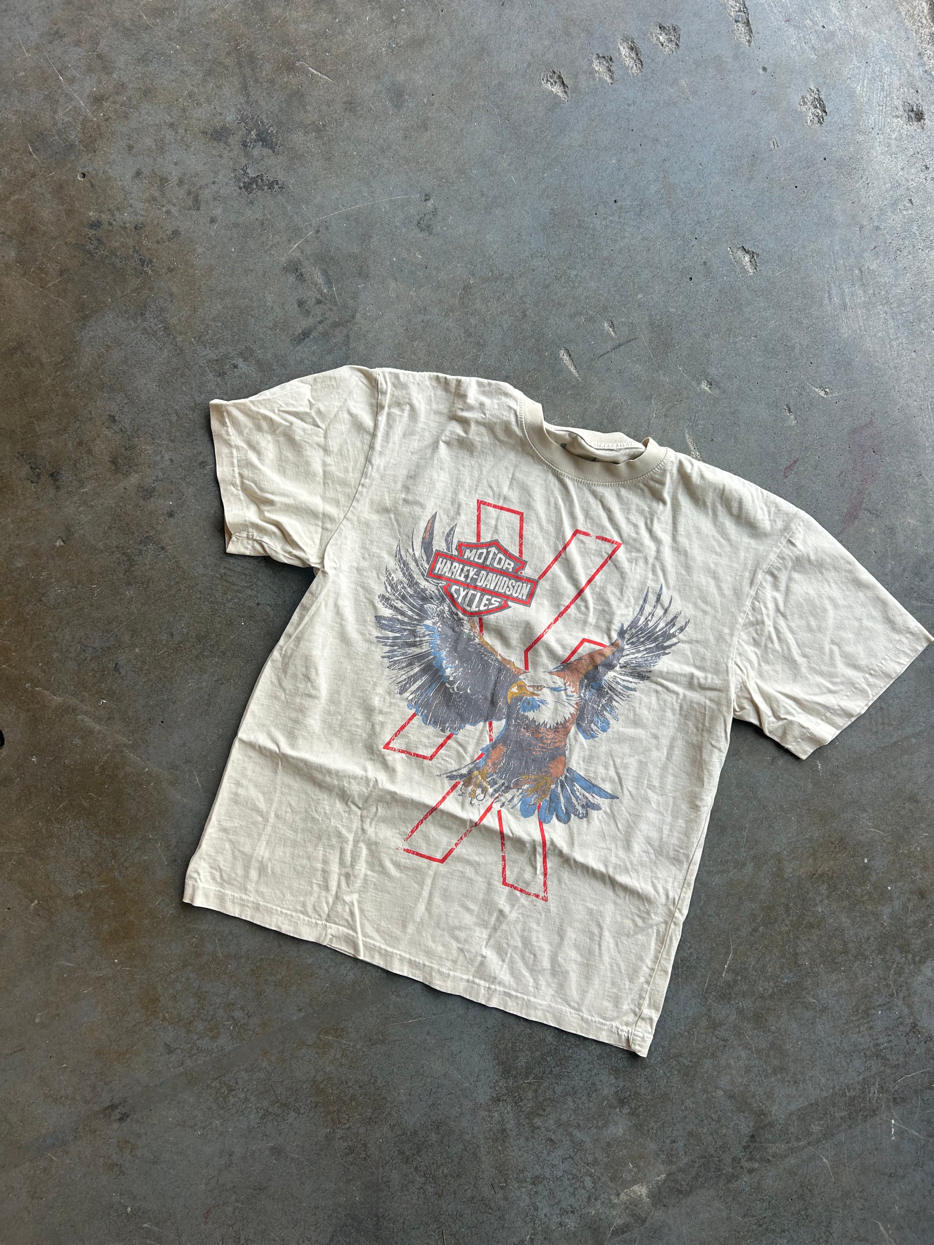 A vintage off-white short-sleeve t-shirt with a distressed graphic of a bird in flight over a red 'X' and the words 'Harley Davidson', laid flat on a textured gray concrete floor