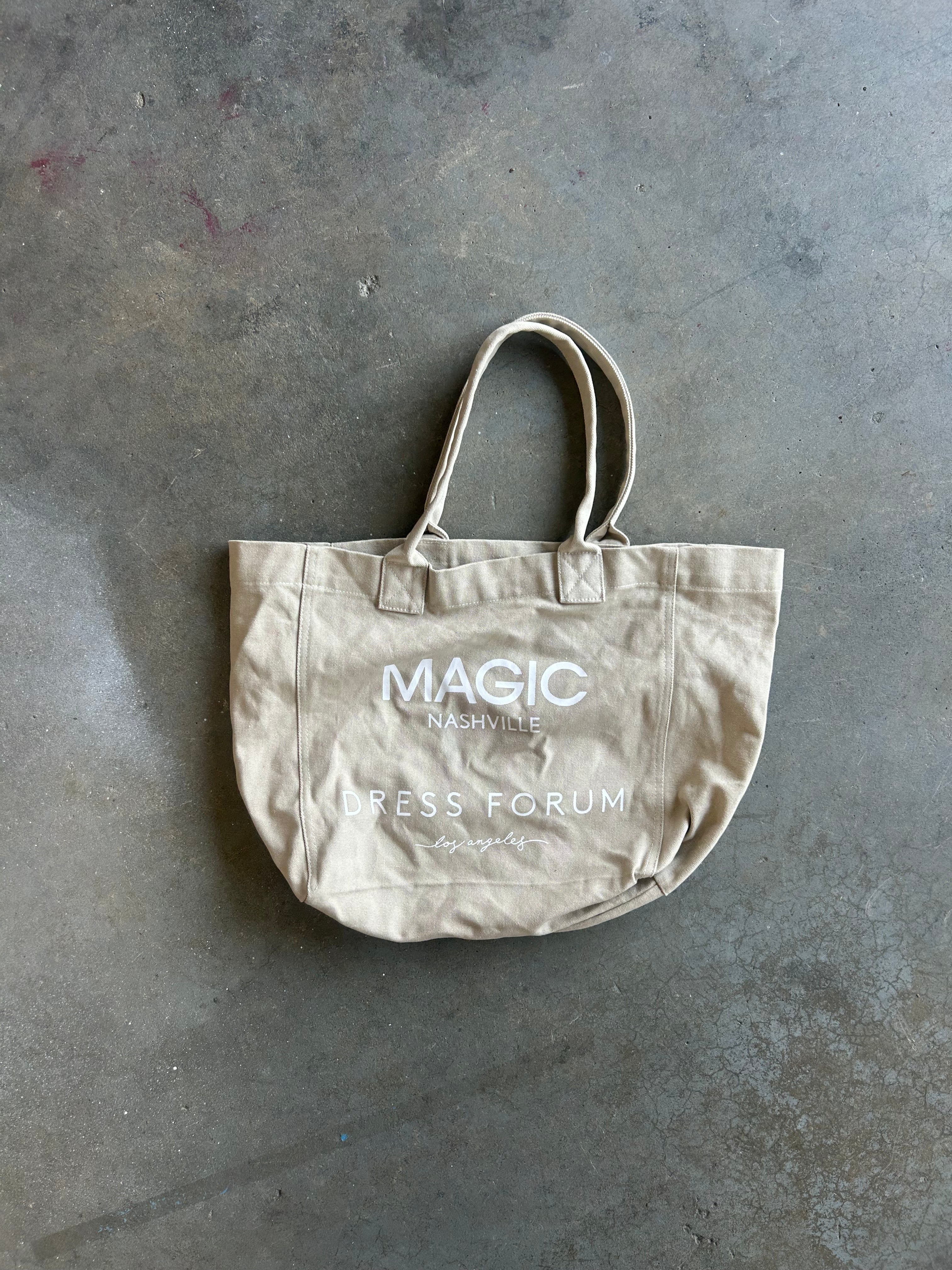 A beige canvas tote bag with white printed text reading 'MAGIC MAGAZINE DRESS FORUM' and 'Original' lies on a textured gray concrete floor