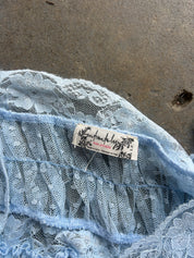 Close-up of a light blue lace garment with a visible sewn-in label reading 'Anchors Aweigh' and 'Made in Japan', laid on a gray concrete surface