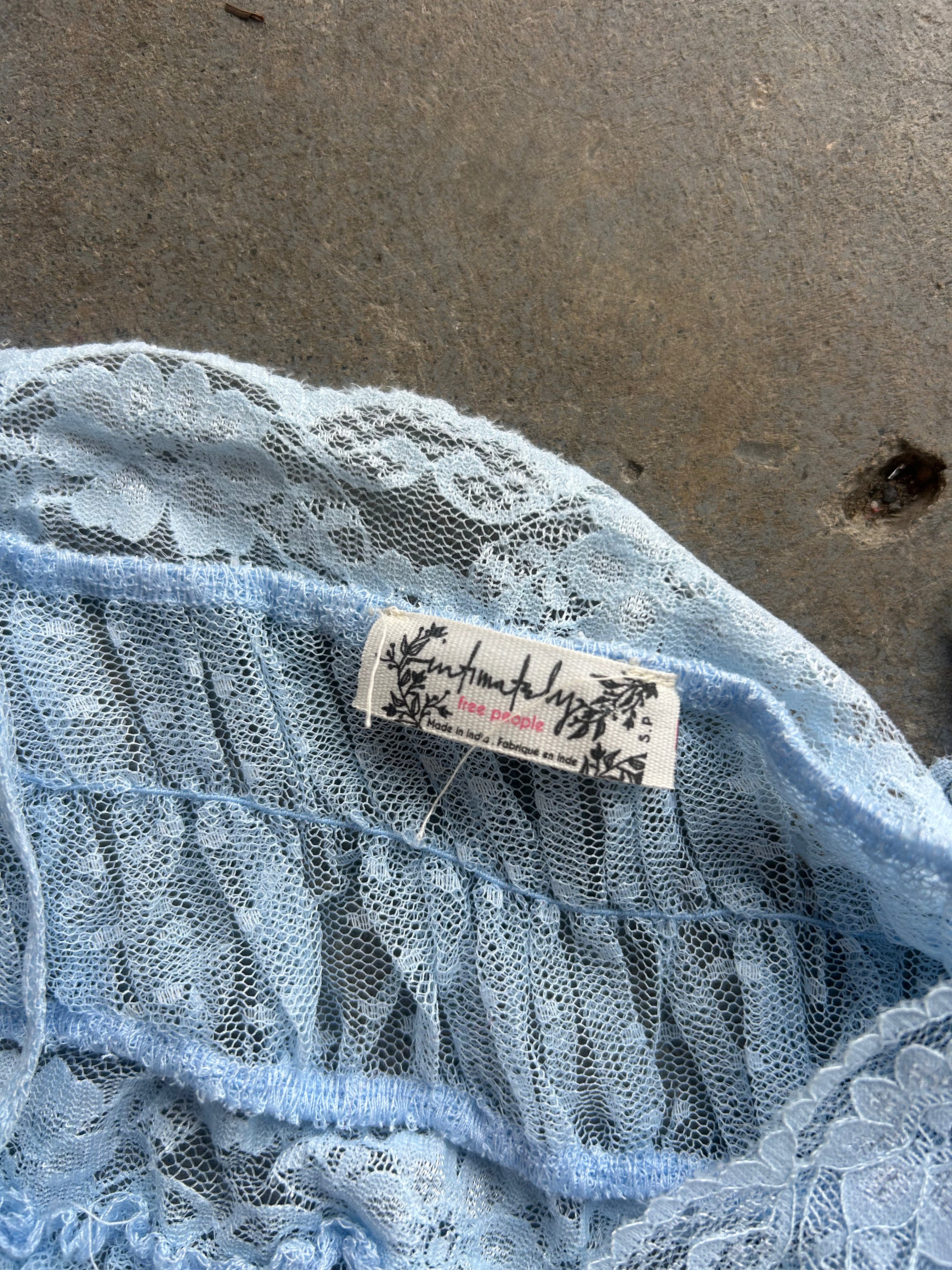Close-up of a light blue lace garment with a visible sewn-in label reading 'Anchors Aweigh' and 'Made in Japan', laid on a gray concrete surface