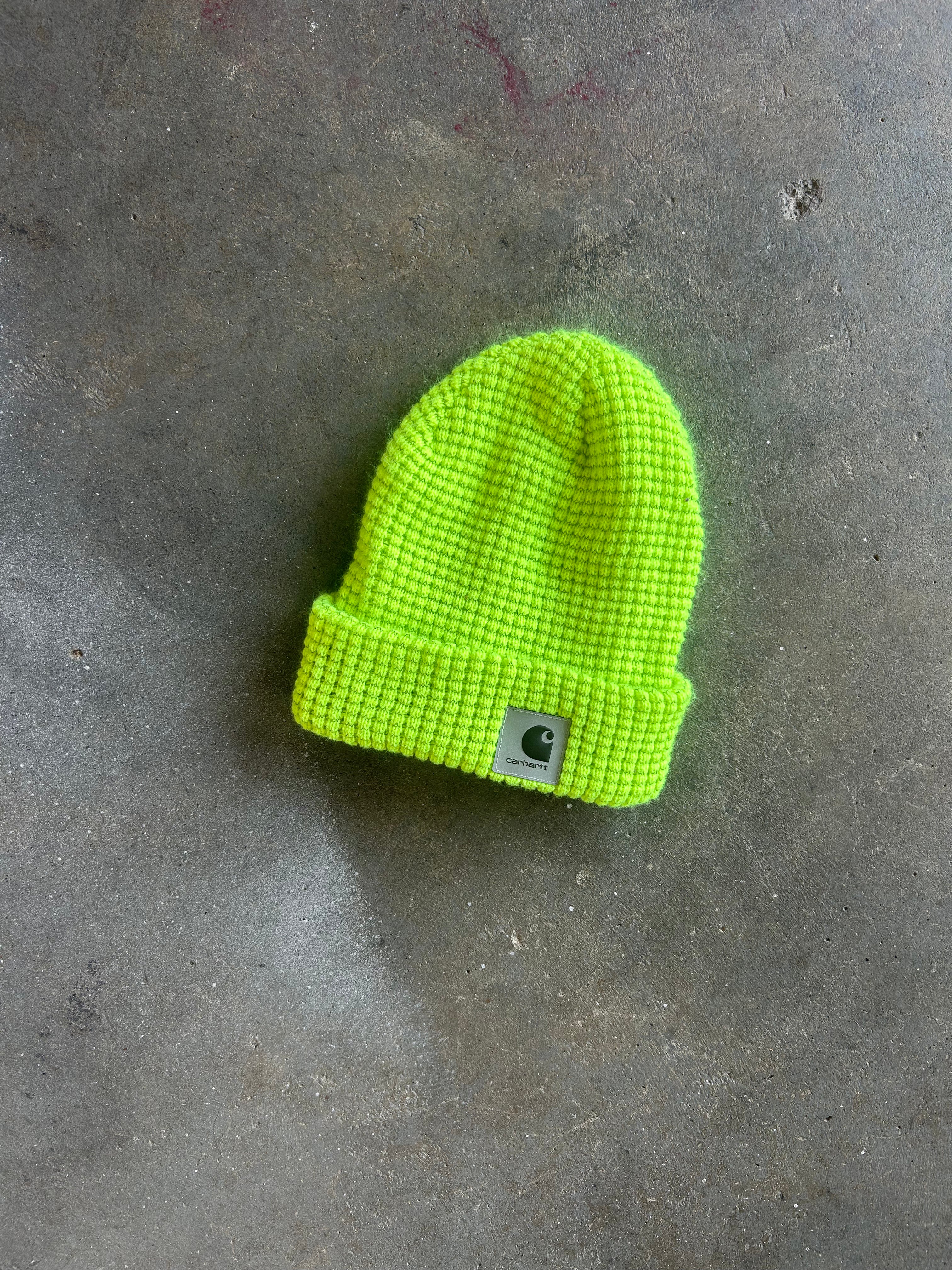 A bright neon yellow-green knit beanie hat with a folded cuff and a small square Carhartt logo patch on the front, placed on a textured gray concrete surface