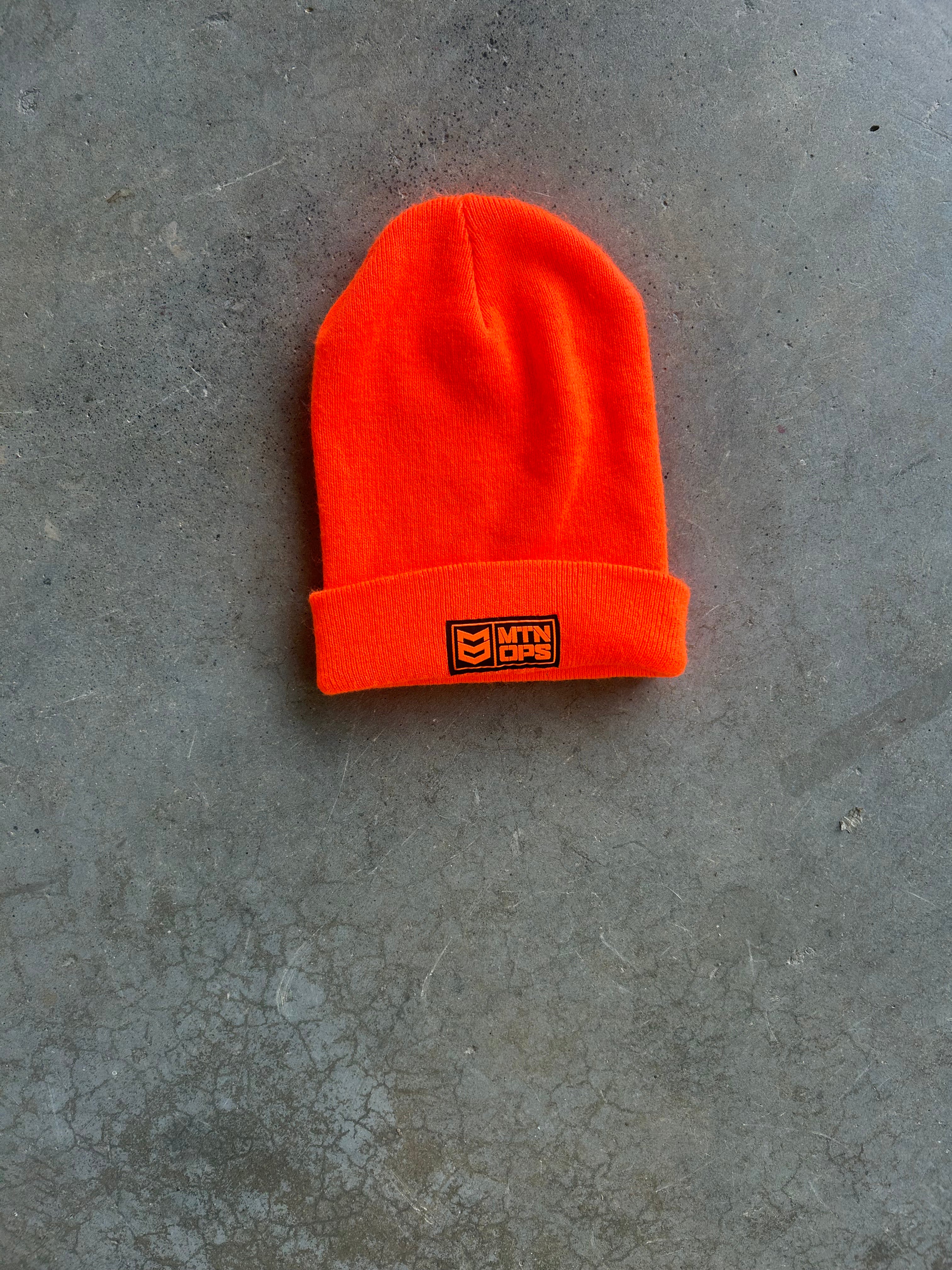 A bright neon orange beanie hat with a folded cuff, featuring a small black embroidered logo patch on the front, laid flat on a textured gray concrete surface