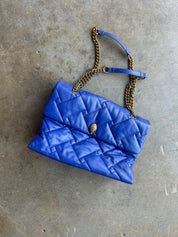 A bright blue quilted leather shoulder bag with a gold chain strap and a small gold clasp, laid flat on a textured gray concrete surface