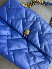 A close-up of a blue quilted leather shoulder bag with a gold chain strap and a gold snake-head clasp, resting on a gray surface