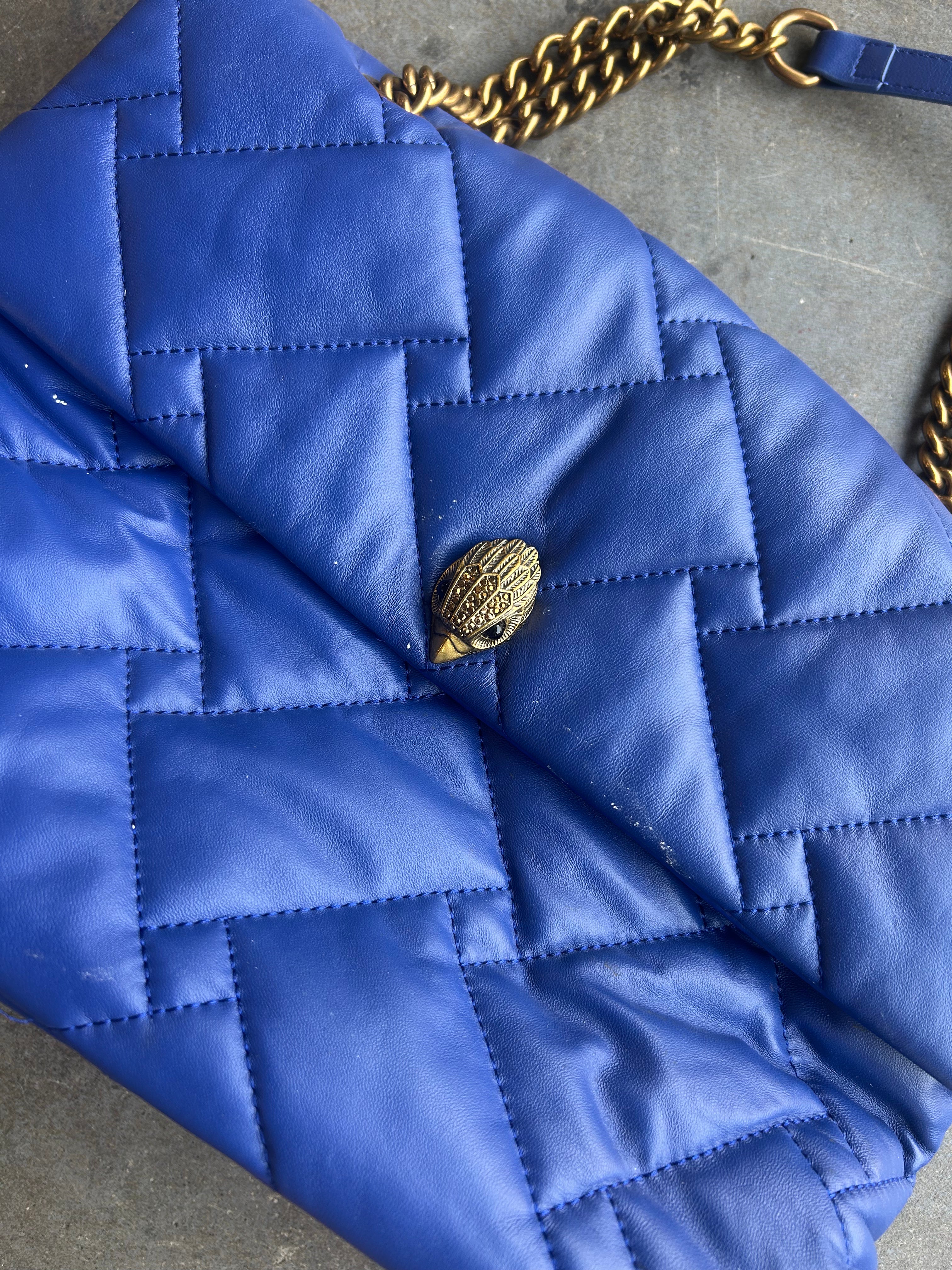 A close-up of a blue quilted leather shoulder bag with a gold chain strap and a gold snake-head clasp, resting on a gray surface