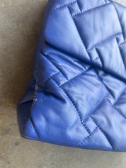A close-up of a blue quilted faux-leather or leather bag corner showing the stitching and texture, resting on a gray concrete floor