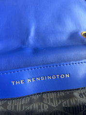 Close-up of a blue leather bag with gold-tone hardware and the text 'THE KENSINGTON' printed in gold on a stitched leather band. A patterned black and gray fabric is visible at the bottom