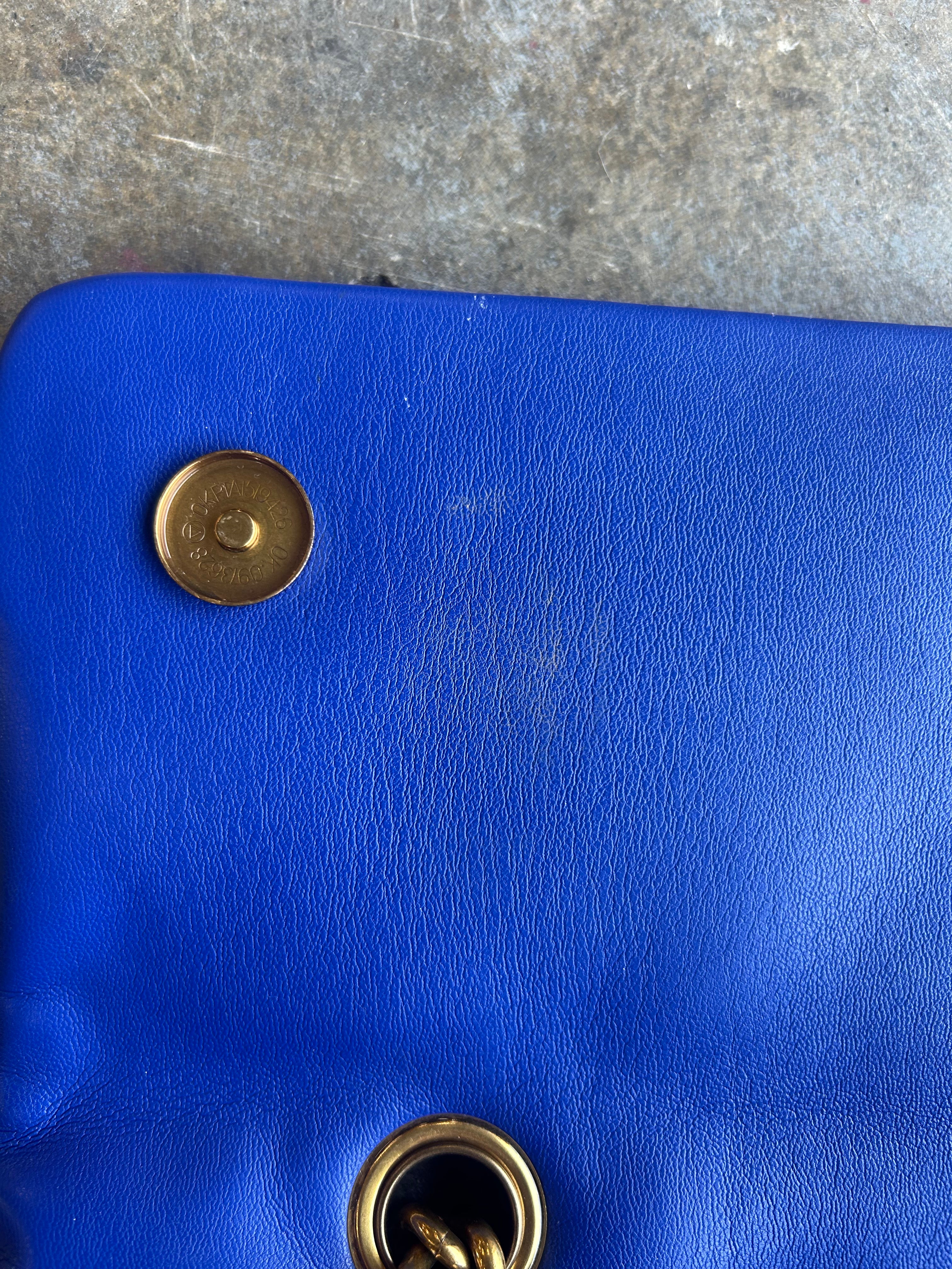 Close-up of a bright blue textured leather bag corner with a gold-tone snap button and metal grommet on a gray stone surface