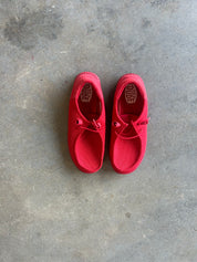 A pair of bright red slip-on shoes with lace-up details, laid flat on a textured gray concrete floor. The brand name 'Hey Dude' is visible on the insole