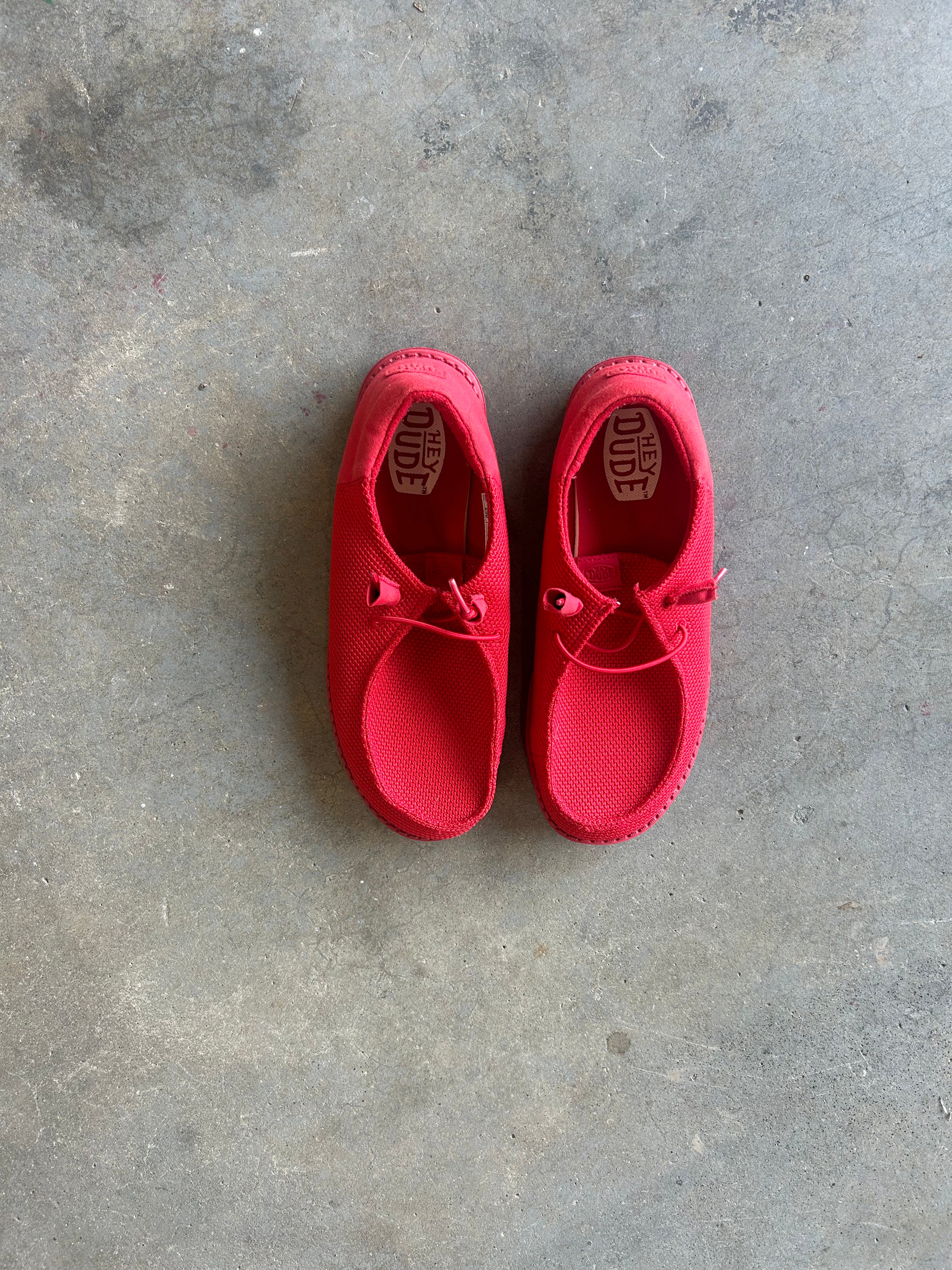 A pair of bright red slip-on shoes with lace-up details, laid flat on a textured gray concrete floor. The brand name 'Hey Dude' is visible on the insole