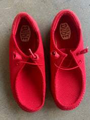 A pair of red slip-on canvas shoes with elastic laces, shown from a top-down view on a gray concrete floor. The brand name 'Hey Dude' is visible on the insoles