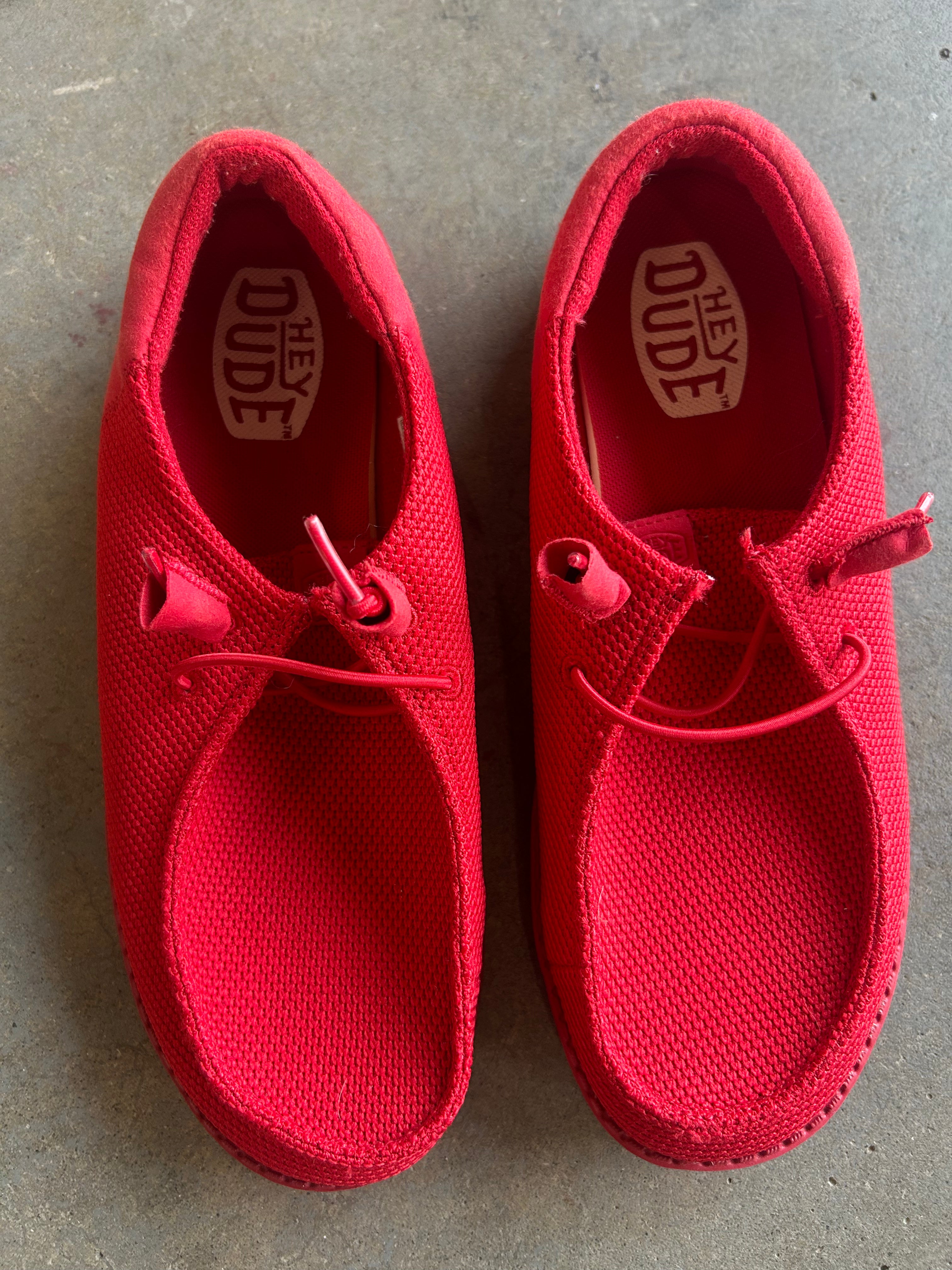 A pair of red slip-on canvas shoes with elastic laces, shown from a top-down view on a gray concrete floor. The brand name 'Hey Dude' is visible on the insoles
