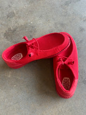 A pair of red canvas slip-on shoes with heart-shaped cutouts and laces, displayed on a gray concrete floor. The brand name 'HEY DUDE' is visible on the insoles