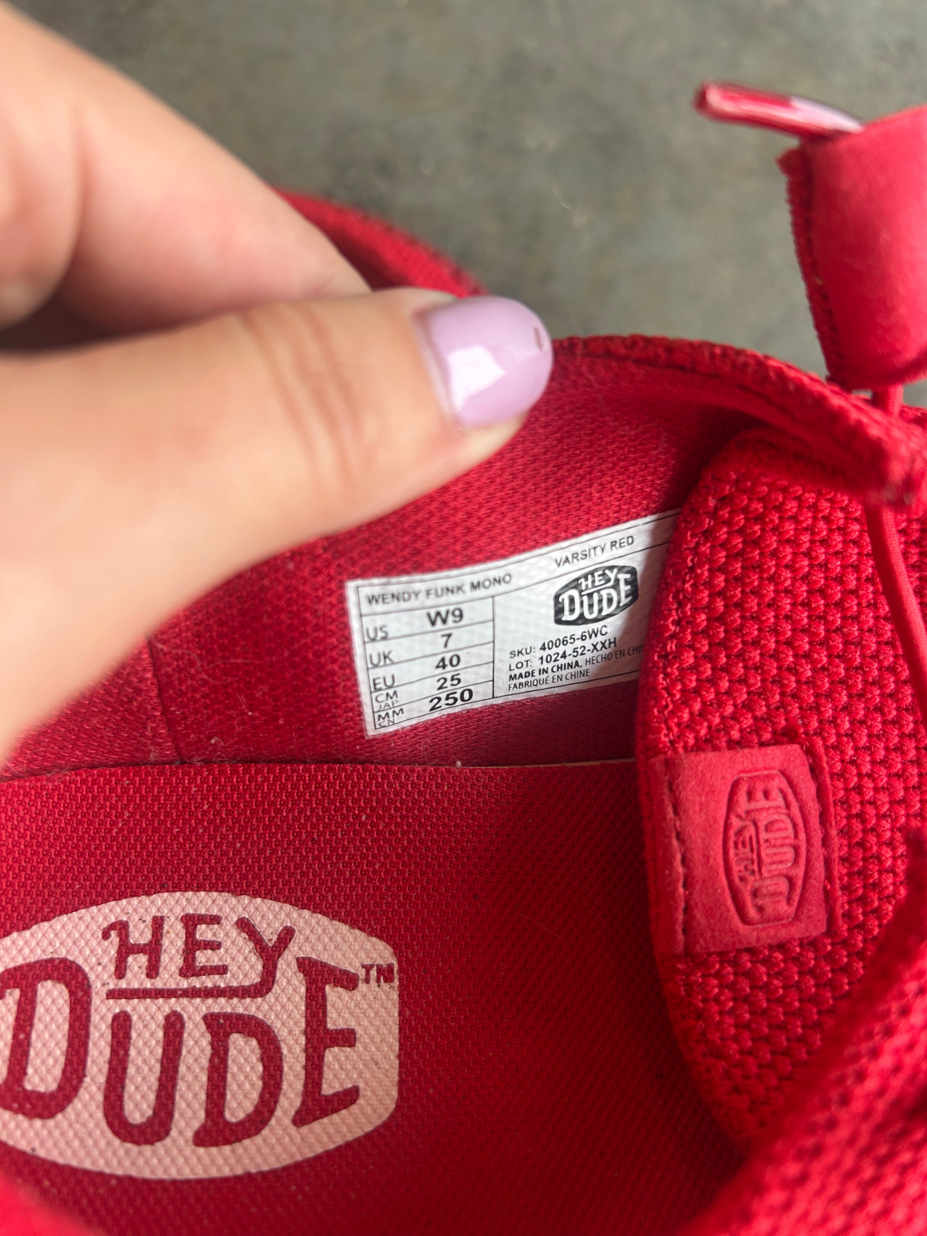 Close-up of a red Hey Dude shoe interior showing the size label and a finger with light purple nail polish touching the inside lining