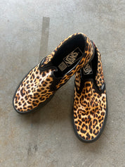 A pair of Vans slip-on sneakers with a leopard print pattern, displayed on a gray concrete floor