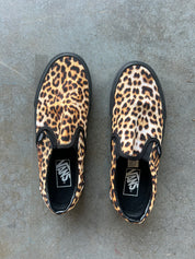 A pair of Vans slip-on sneakers with a leopard print pattern, photographed from a top-down view on a gray concrete floor