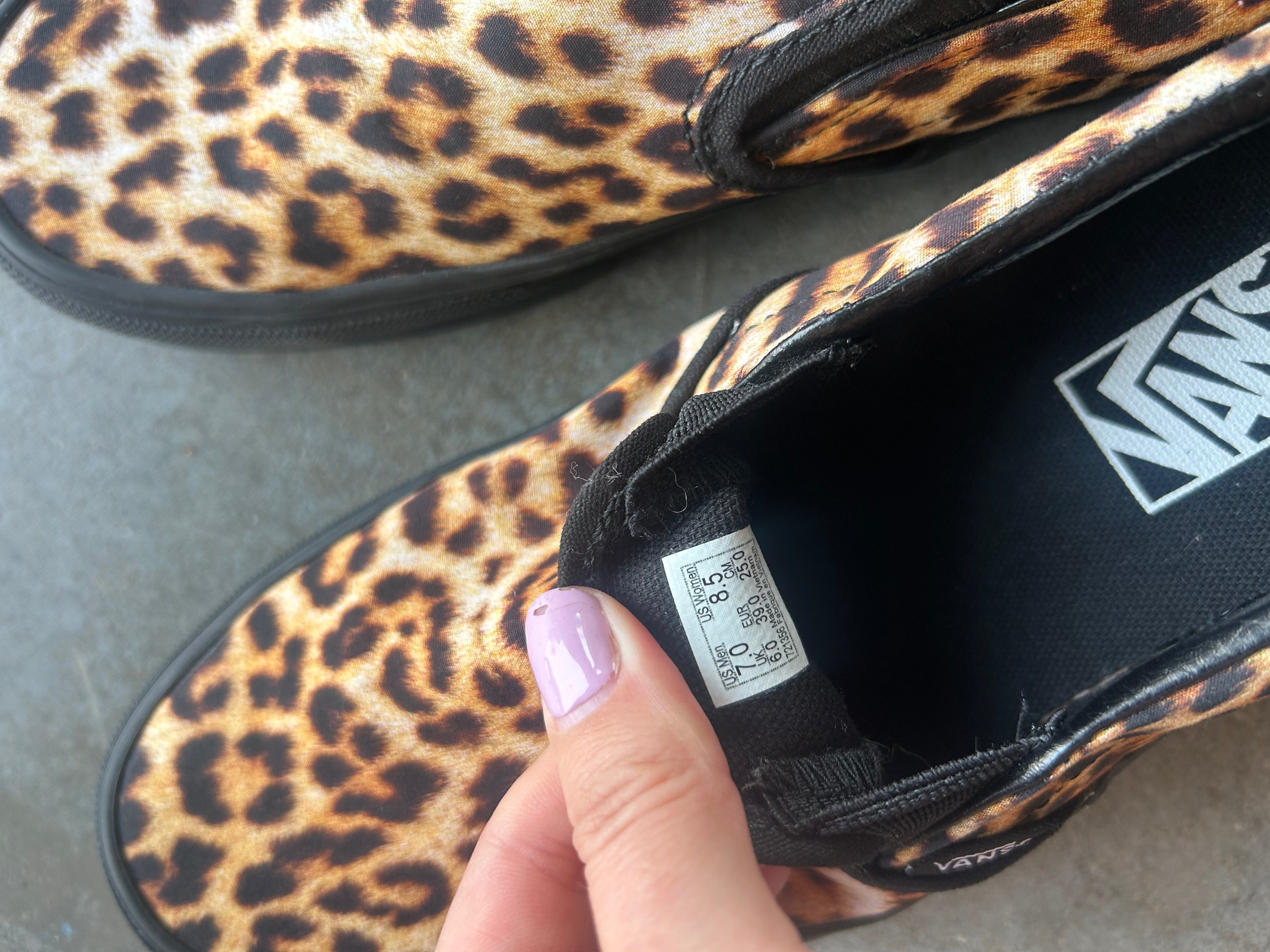 A close-up of a Vans slip-on shoe with a leopard print pattern, held by a hand with lavender nail polish. The shoe has a black interior with the Vans logo visible, and a size tag is shown